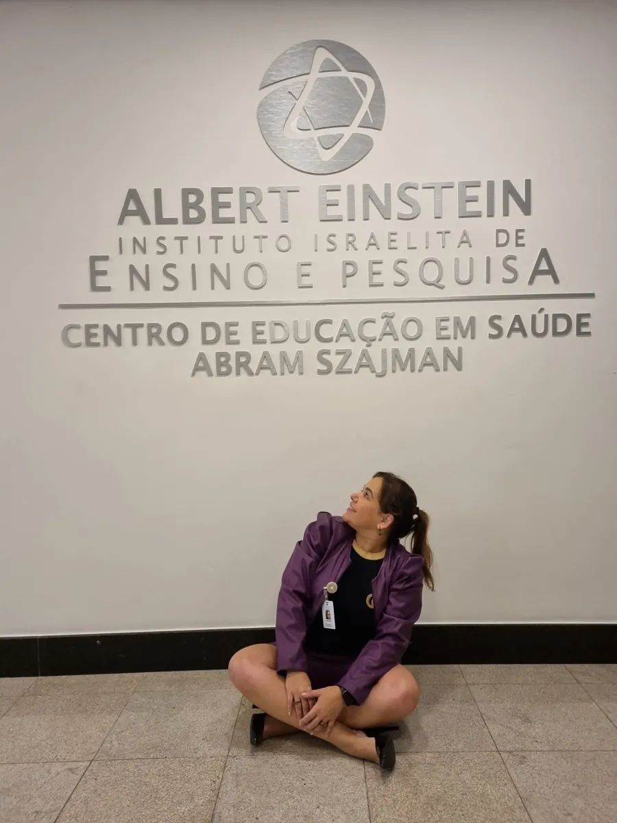 Adriane sentada no chão, sorrindo e olhando para o letreiro do Centro de Educação em Saúde do Instituto Albert Einstein